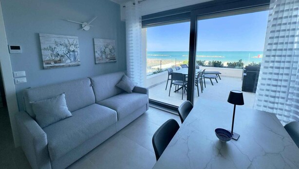 Una sala de estar moderna con un sofá, una mesa de comedor y sillas. Hay dos cuadros en la pared, un ventilador de techo y vistas a la playa a través de amplias ventanas.