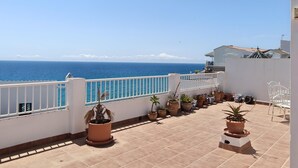 Un balcón con plantas en macetas, barandilla blanca y vista al mar.