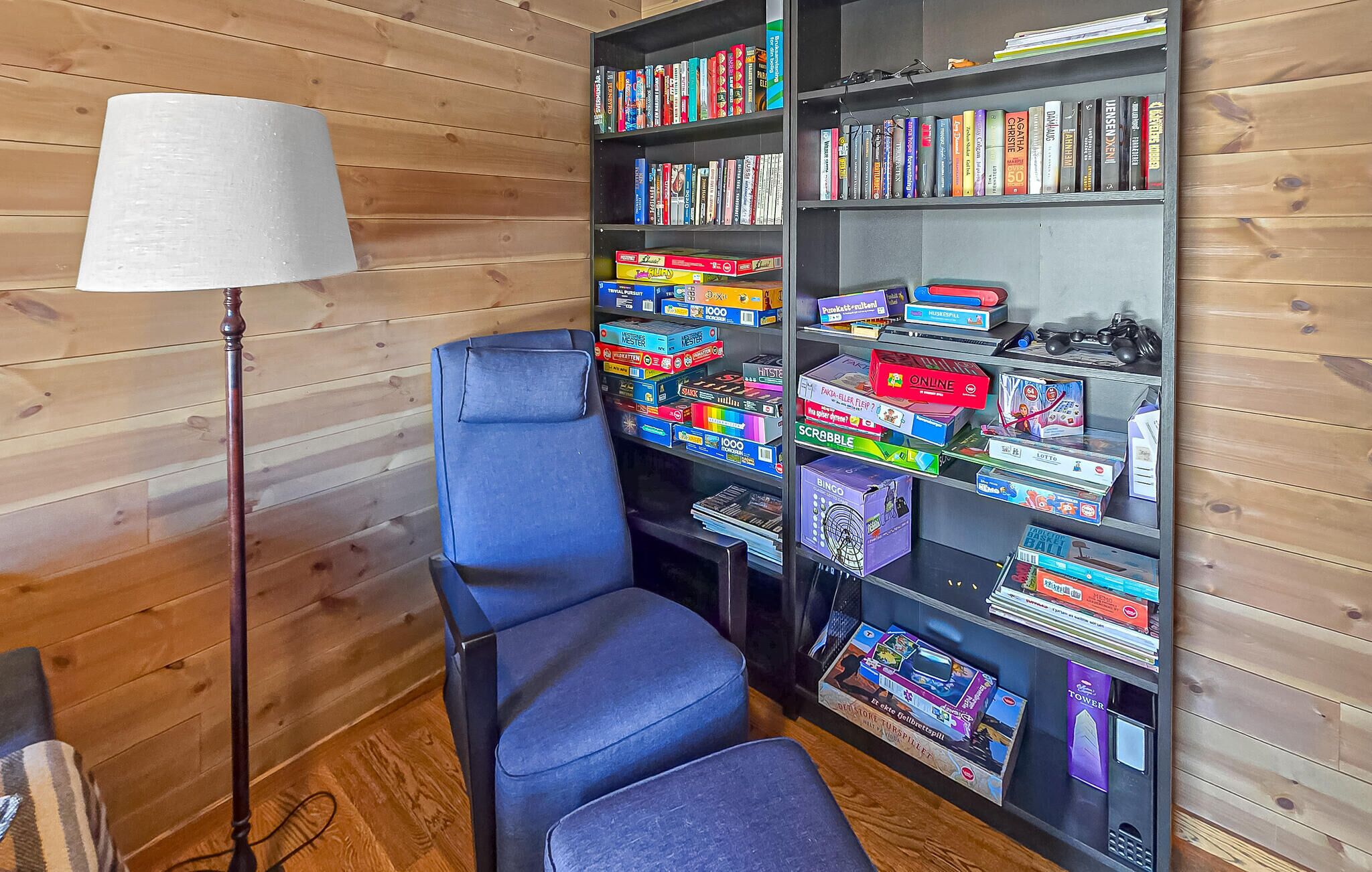 A room with a blue chair, a bookshelf with various board games, and a lamp.