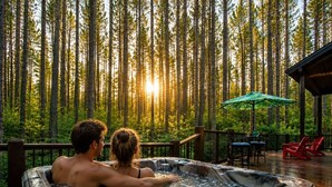 Ein Whirlpool mit zwei Personen, umgeben von einer Terrasse mit Stühlen und einem Sonnenschirm, mit Blick auf einen Wald.