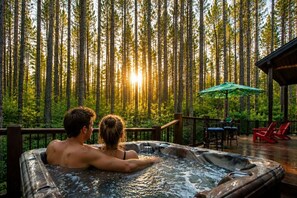 Ein Whirlpool mit zwei Personen, umgeben von einer Terrasse mit Stühlen und einem Sonnenschirm, mit Blick auf einen Wald.
