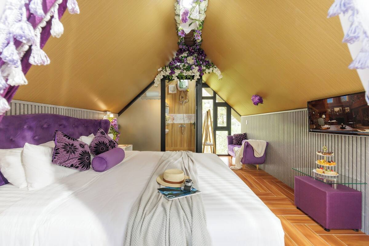 A bedroom with a bed, pillows, a hat, a camera, and a tray table.