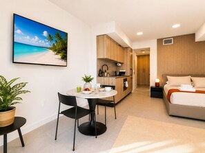 Quarto de hotel moderno com cama, pequena cozinha, mesa de jantar com cadeiras e uma grande janela com vista para a praia.