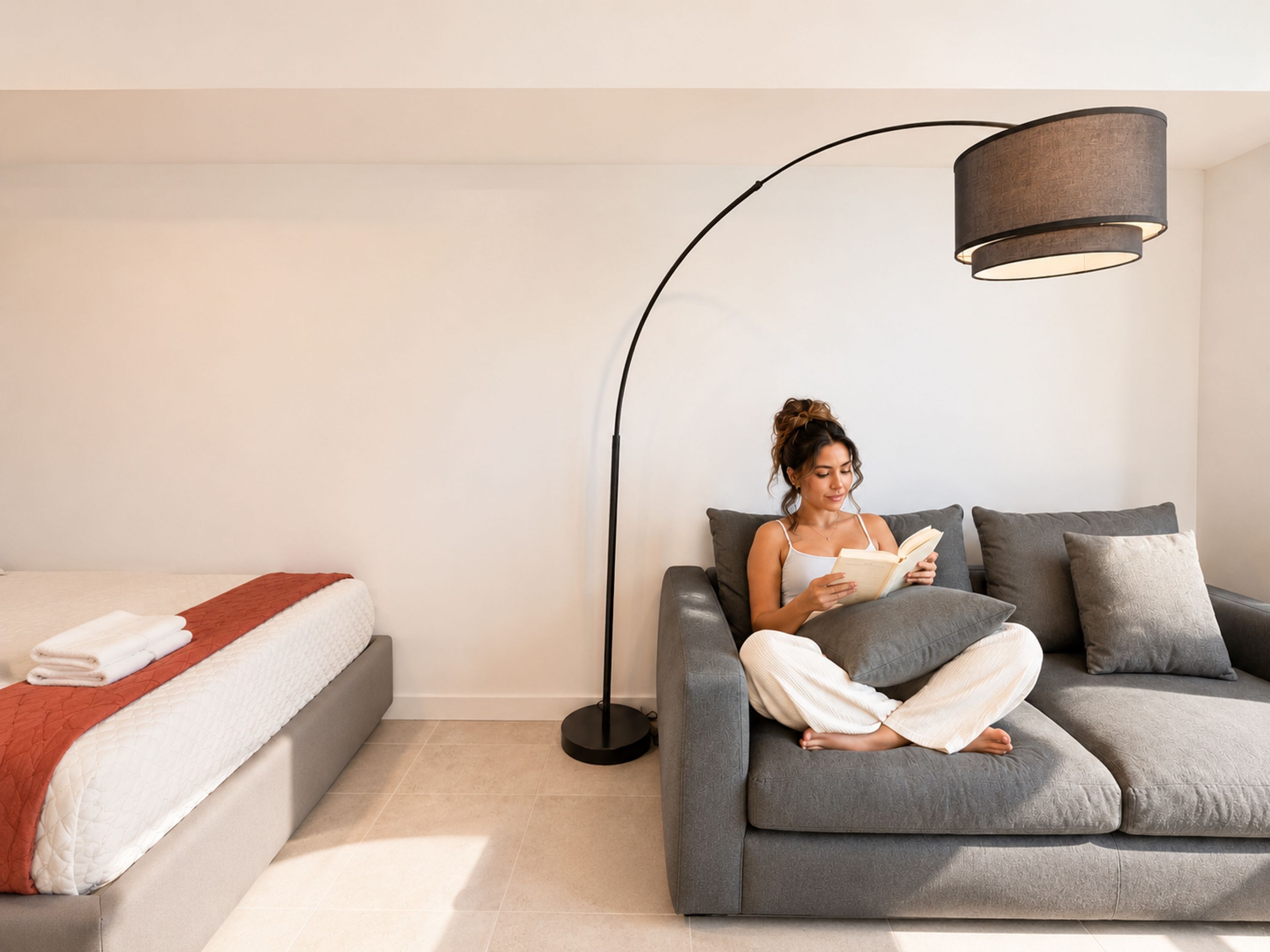 A modern living room with a grey sofa, a floor lamp, a bed with a white and red blanket, and a white wall.