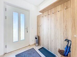 A modern entryway with a white door, a wooden wall with hooks, a trash can, a mat, and a basket with an umbrella. - Apartment Maria by Interhome (Großklein)