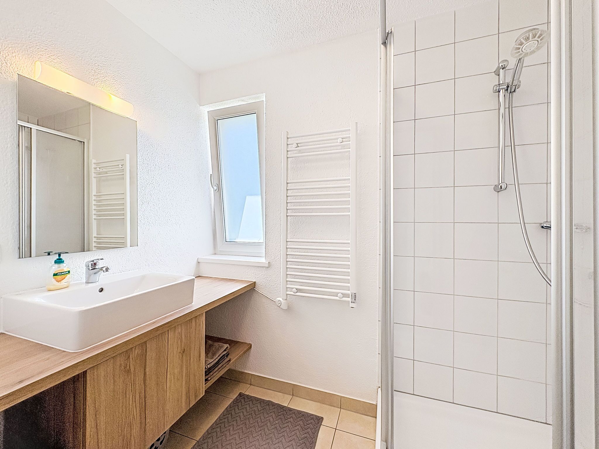 A bathroom with a white sink, mirror, window, towel rack, and shower.