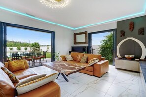 Ein modernes Wohnzimmer mit einer Couch, einem Couchtisch und einem Fernseher, großen Fenstern und einem Essbereich mit Stühlen.
