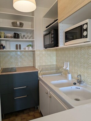 A kitchen with a microwave, sink, and shelves with kitchenware. - Le Portique, location T2 pour 4 personnes à Aiguebelle, vue mer, 350m plage (LE LAVANDOU)