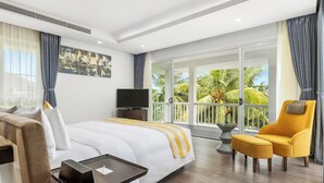 Habitación de hotel con cama, televisor, balcón y vista a palmeras, además de un sillón amarillo.