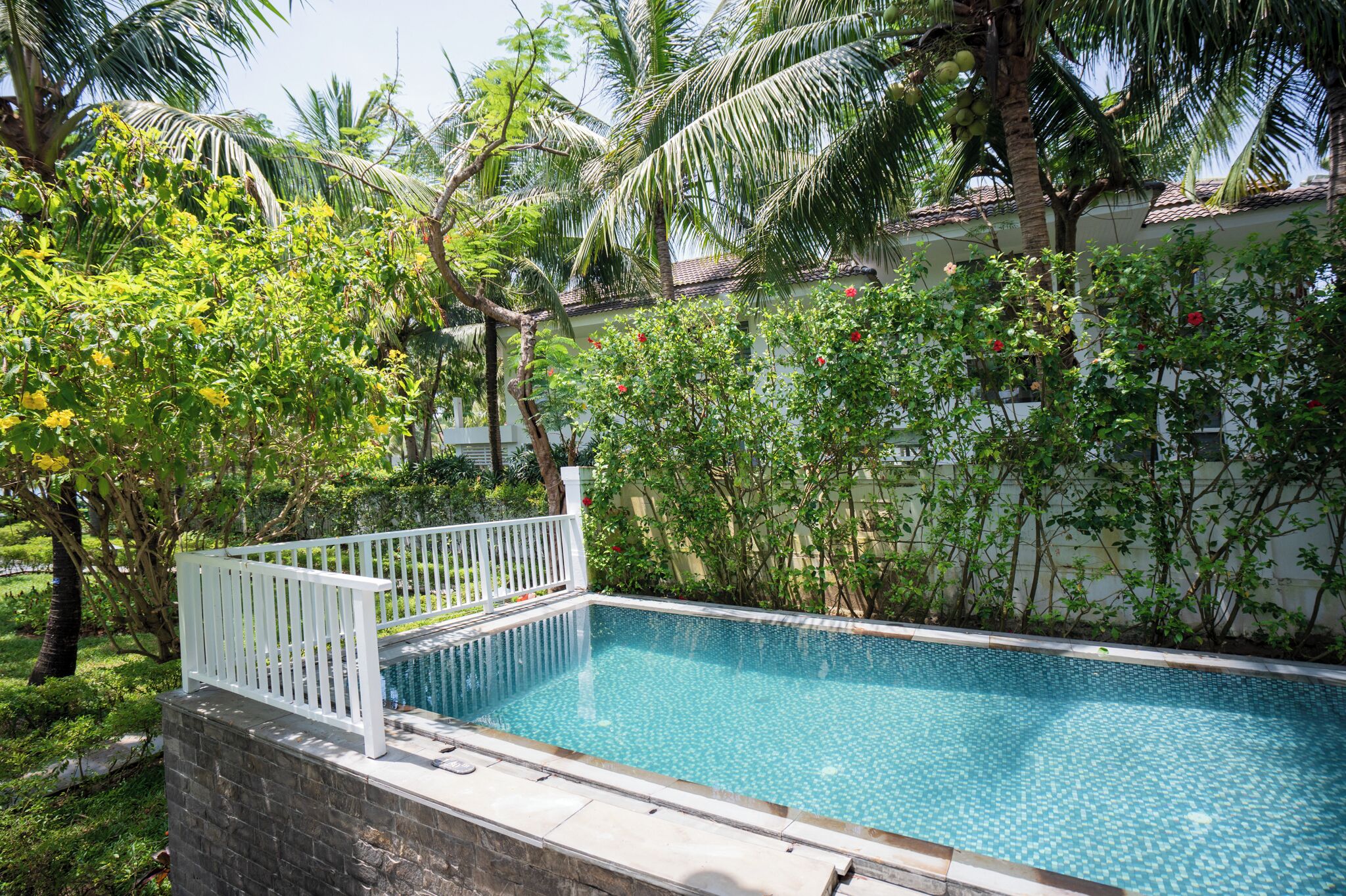 Ein rechteckiger Swimmingpool mit weißem Zaun, umgeben von üppiger Vegetation und Palmen.