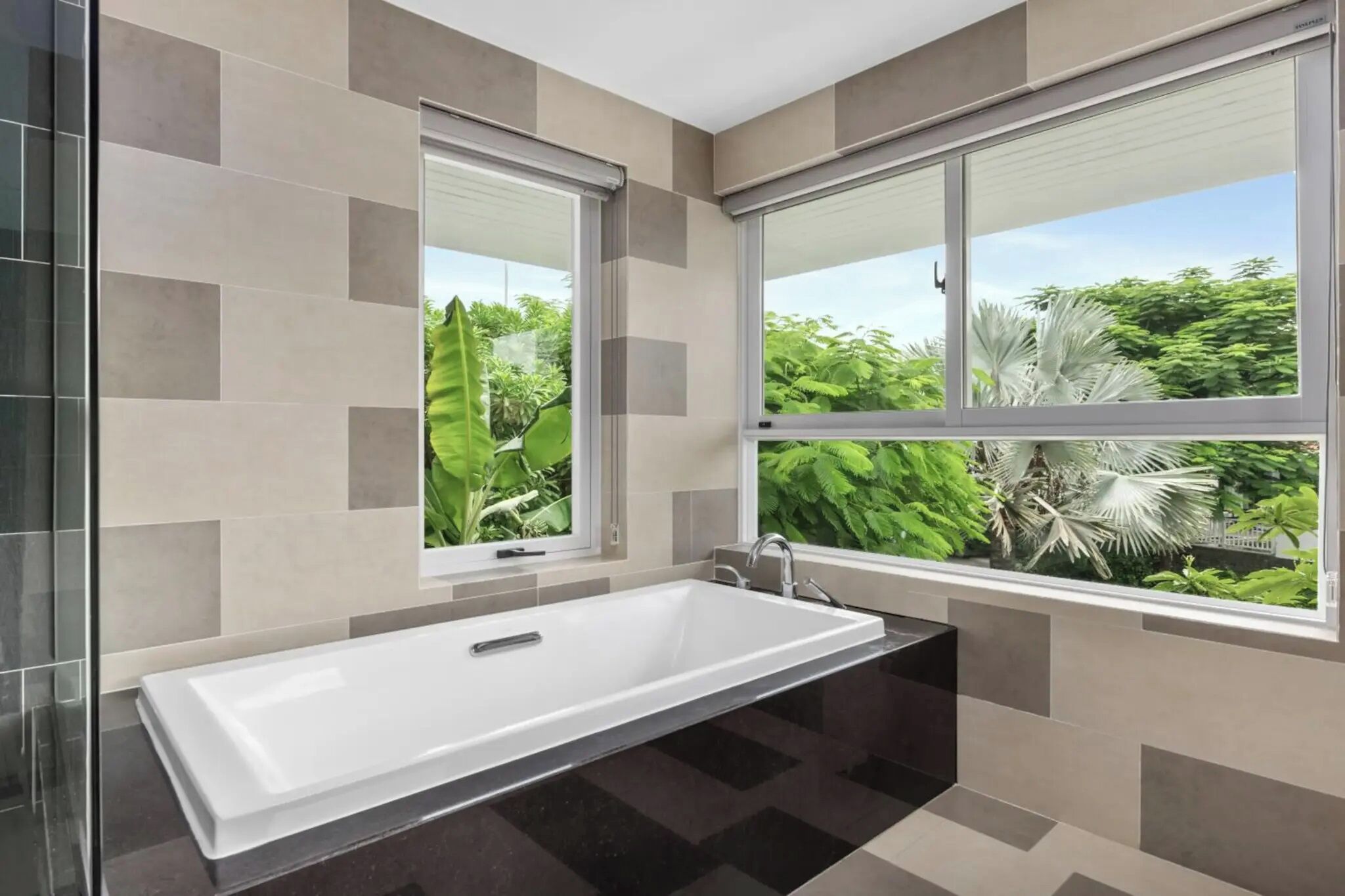 A modern bathroom with a large bathtub, two windows, and a view of greenery outside.