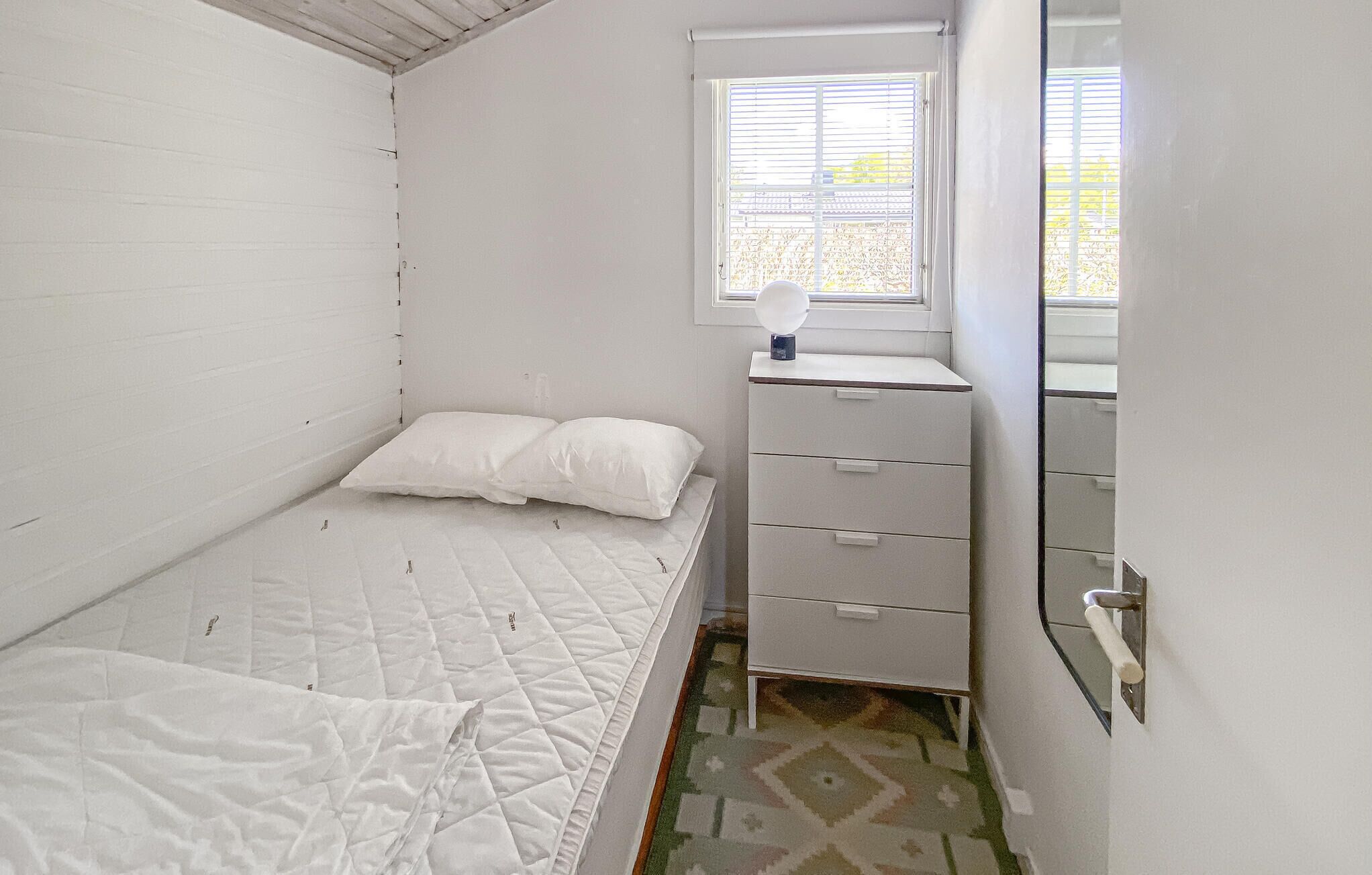 A small, white room with a single bed, a dresser with drawers, a mirror, and a window with blinds.