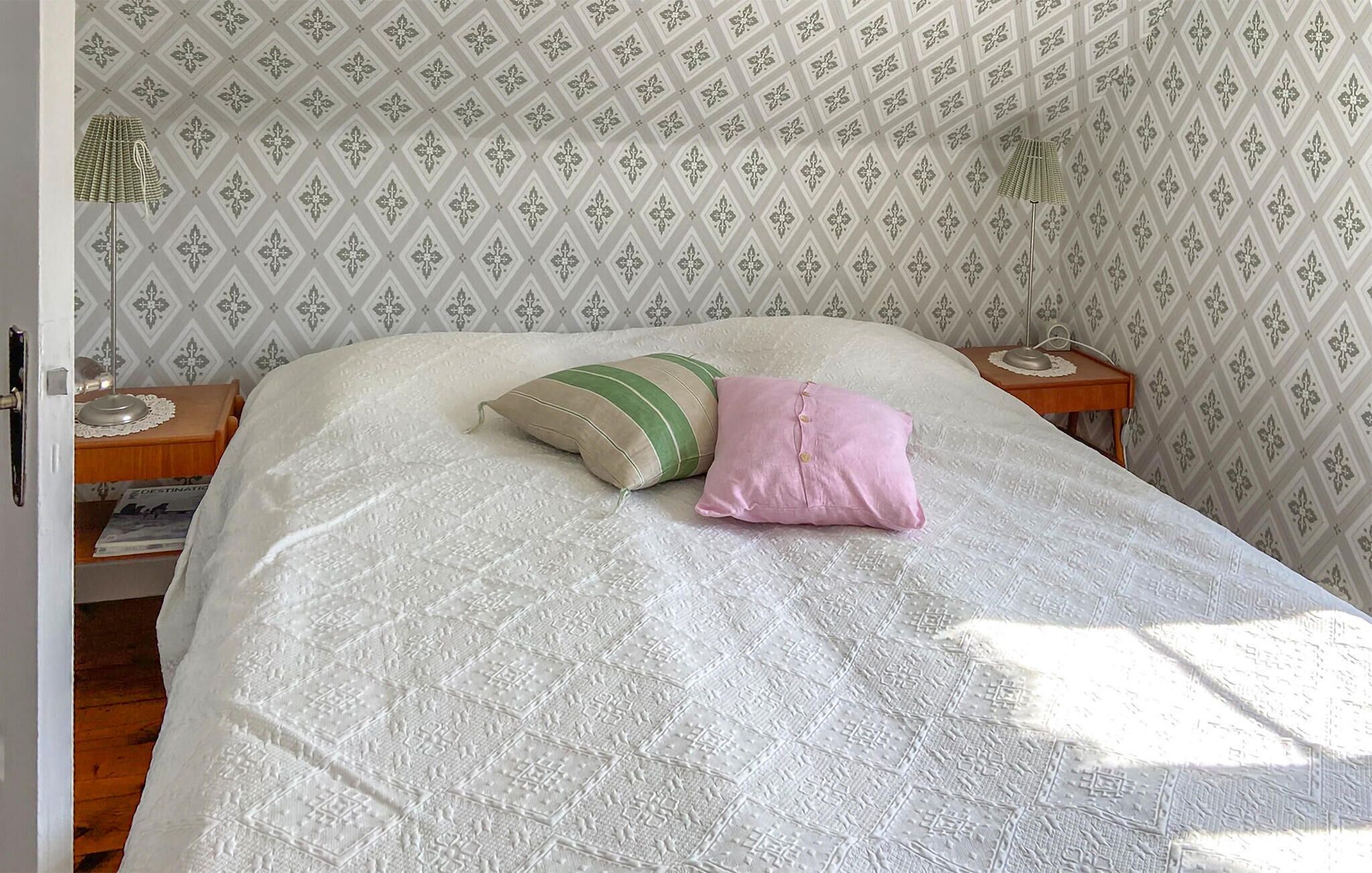 A bed with a white bedspread, two pillows, a nightstand with a lamp, and a patterned wallpaper background.
