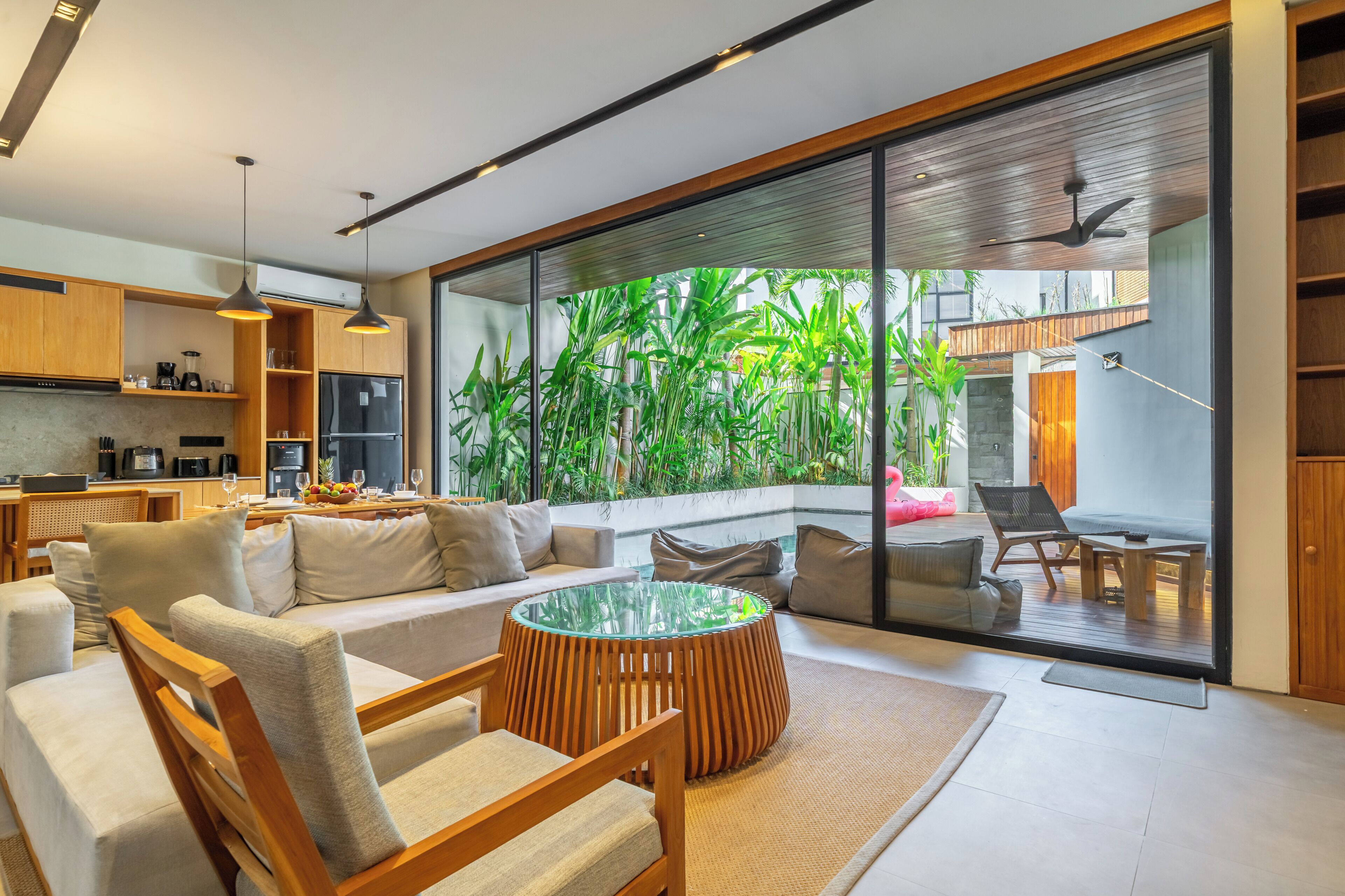 A modern living room with a glass coffee table, wooden furniture, and a view of a lush garden through large windows.