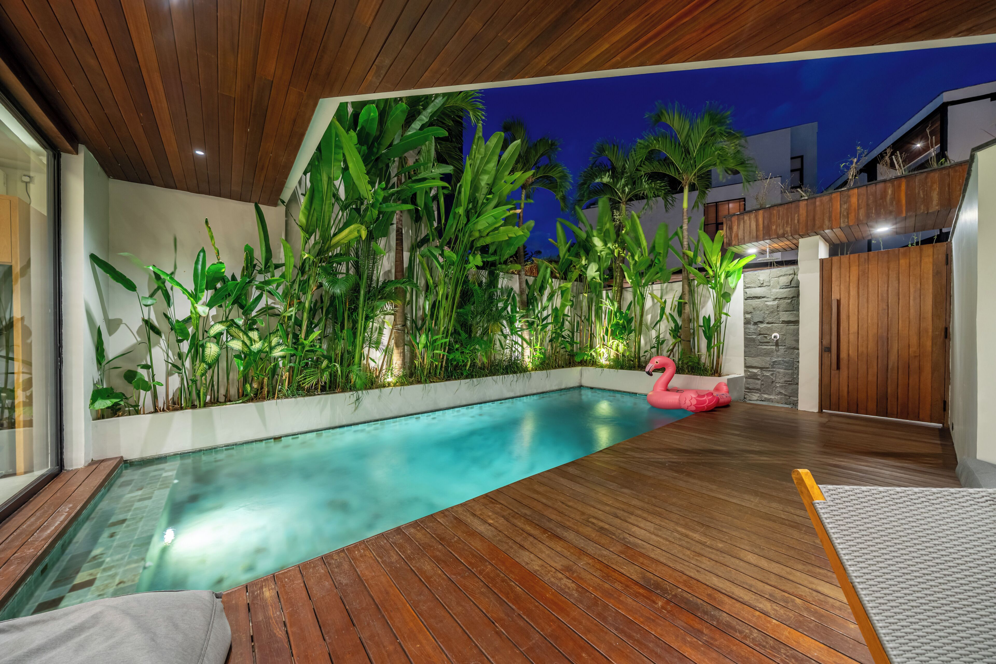 Una piscina con bordo in legno, circondata da piante tropicali rigogliose e un gonfiabile a forma di fenicottero rosa.