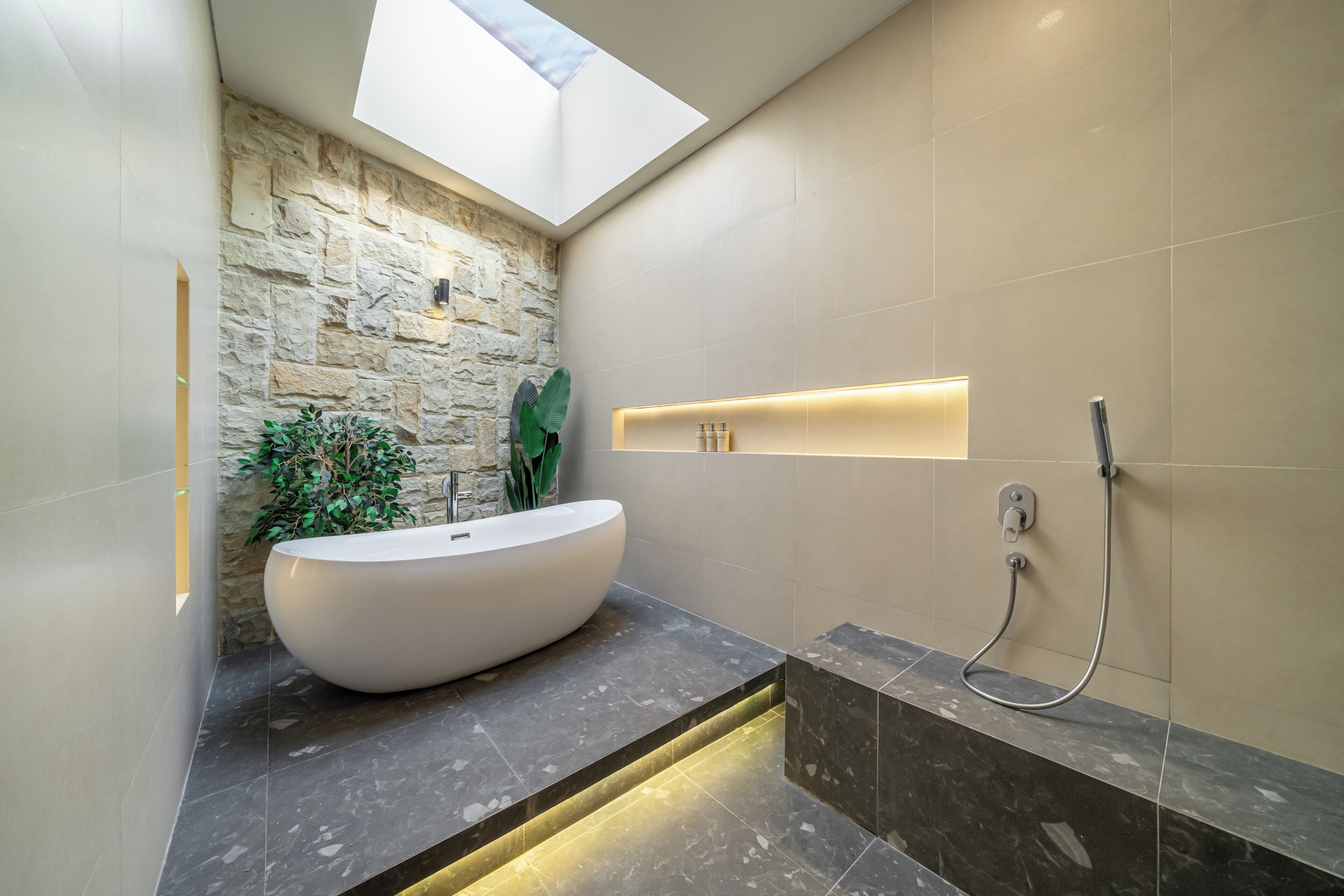 A modern bathroom with a freestanding bathtub, a shower, a skylight, and a stone accent wall.
