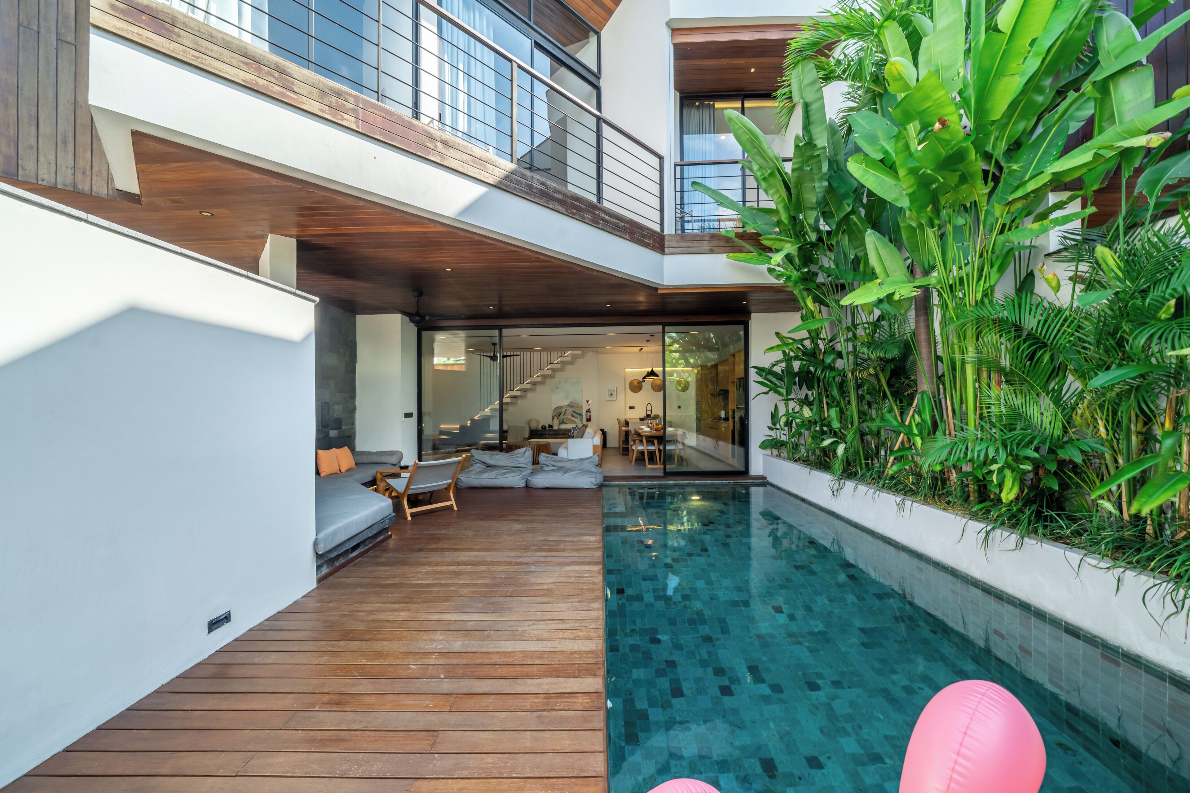 A modern indoor pool area with wooden decking, a seating area with cushions, and lush greenery.
