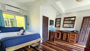 Una habitación con cama, piso de madera, una cocina con armarios y una ventana con cortinas.