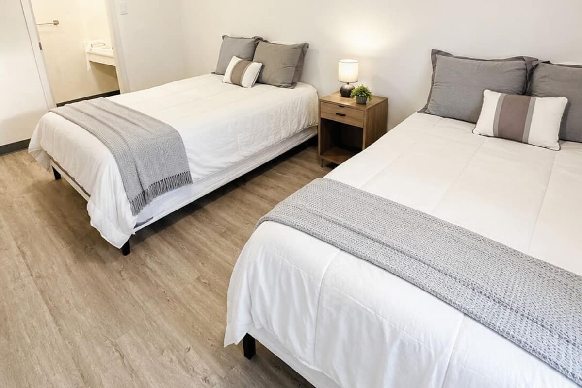 A hotel room with two beds, a bedside table with a lamp, and a door leading to a bathroom.
