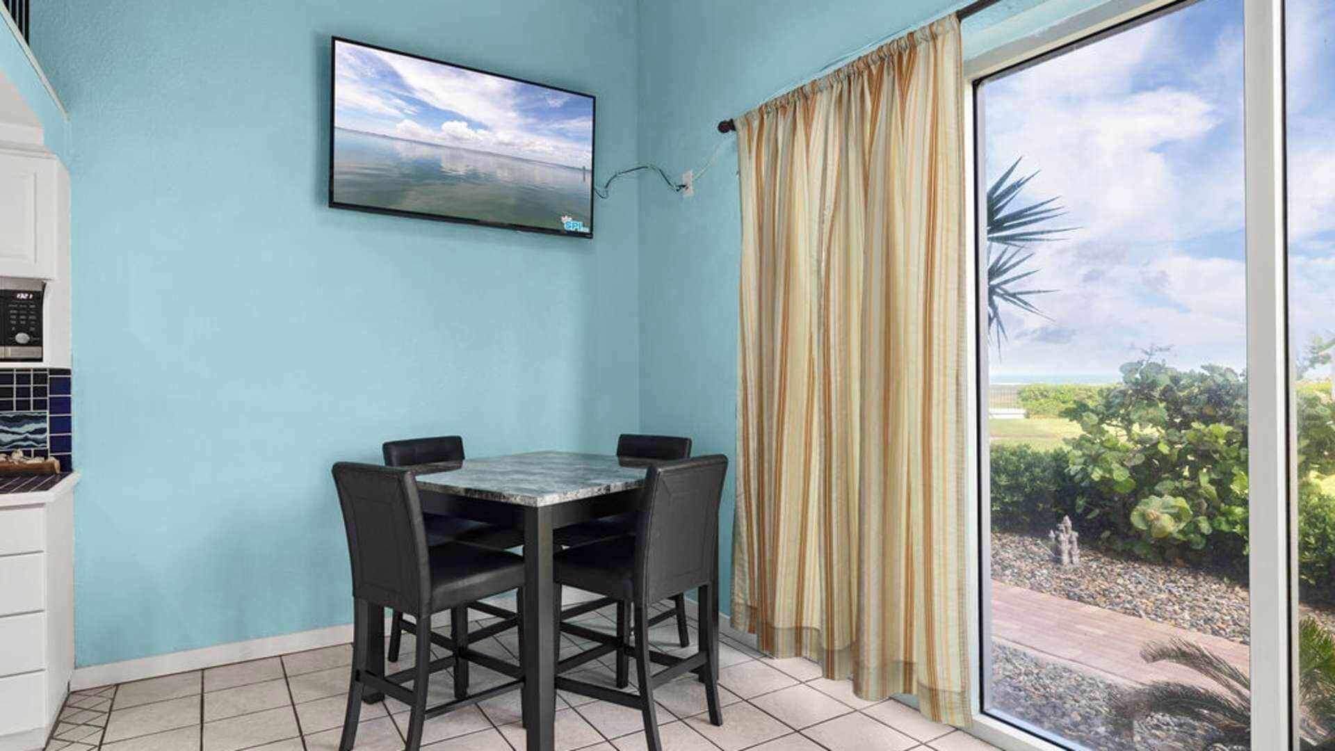 A dining area with a table and chairs, a painting on the wall, and a view through a window with curtains.