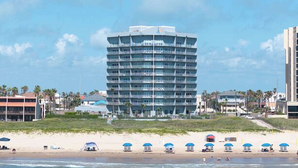 Hotel de varios pisos con balcones, situado cerca de una playa con sillas de tomar el sol y sombrillas.