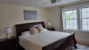 Une chambre à coucher comprenant un lit, deux tables de chevet, un ventilateur de plafond, une fenêtre avec des rideaux et un tableau accroché au mur.