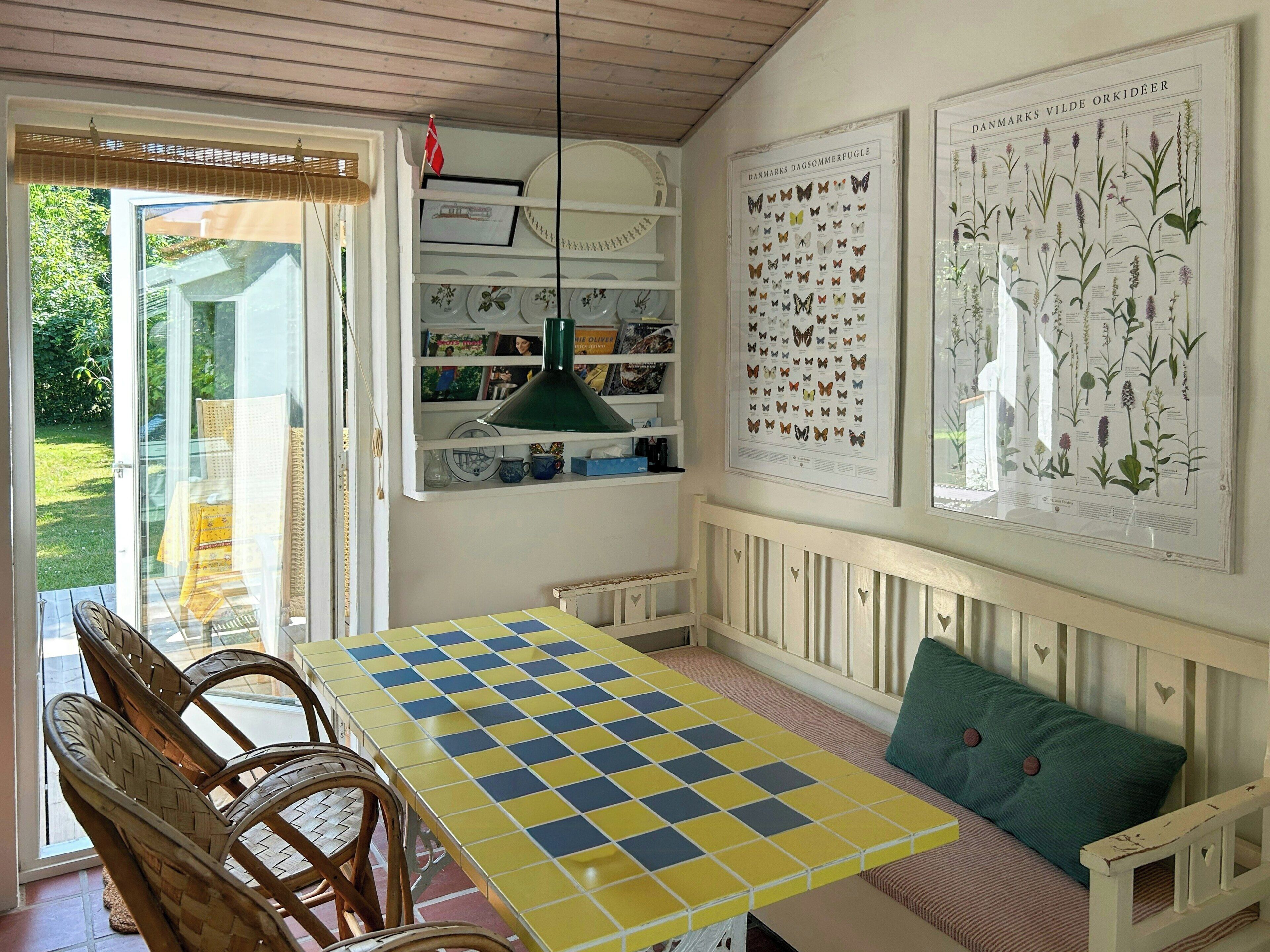 Un comedor con una mesa a cuadros, sillas de mimbre, un banco con recortes en forma de corazón e impresiones botánicas en la pared.