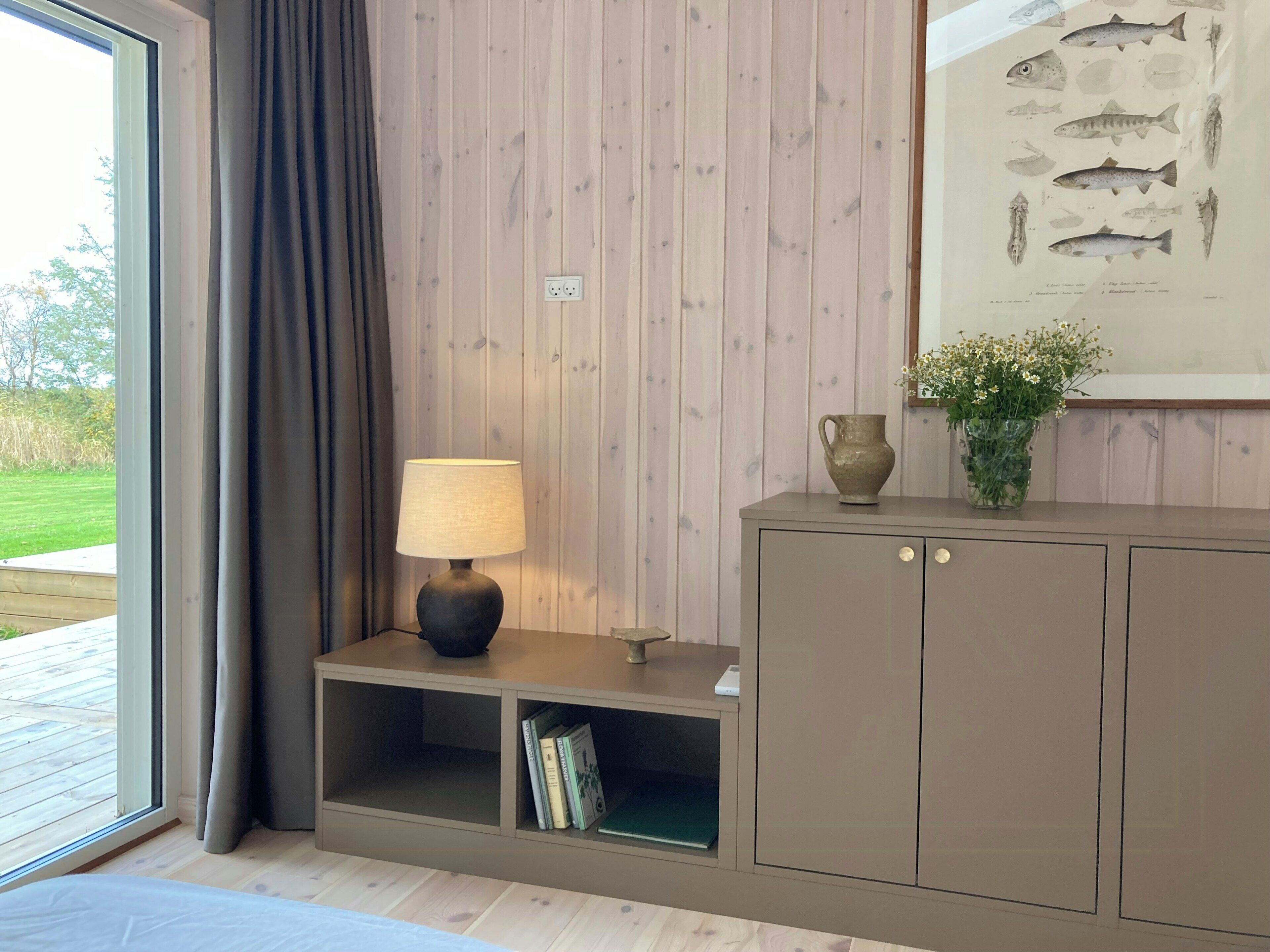 A room with a wooden paneled wall, a lamp on a side table, a cabinet with a vase of flowers, and a framed fish illustration.
