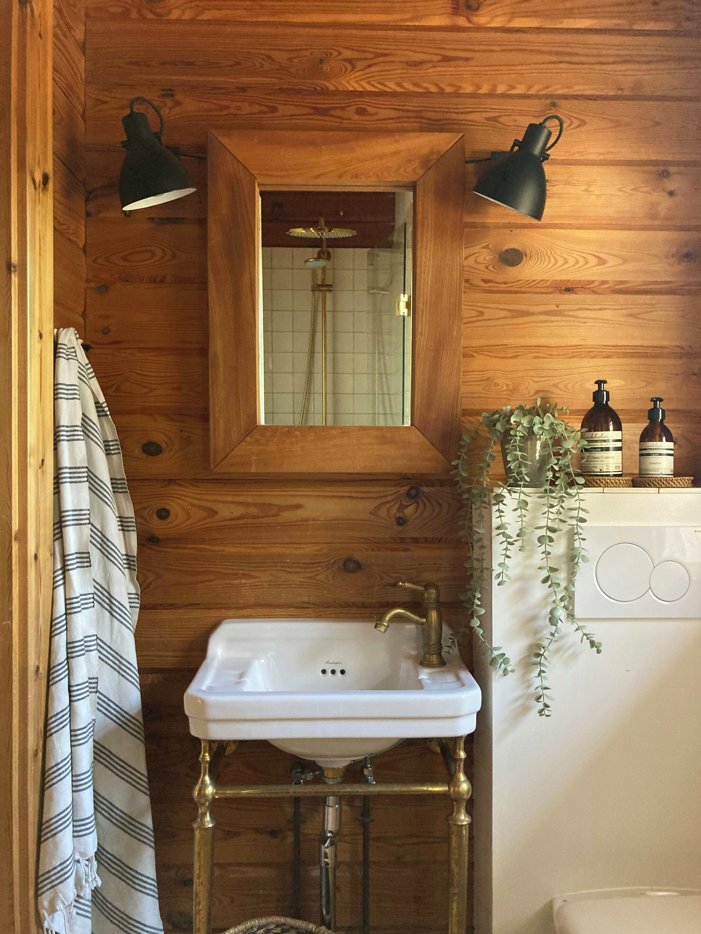 A wooden cabin bathroom with a white sink, two wall-mounted lamps, a mirror, and a shower.