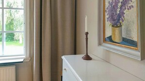 Ein Zimmer mit Fenster, einem weißen Schminktisch, einem gerahmten Gemälde und einer Kerze auf dem Schminktisch.