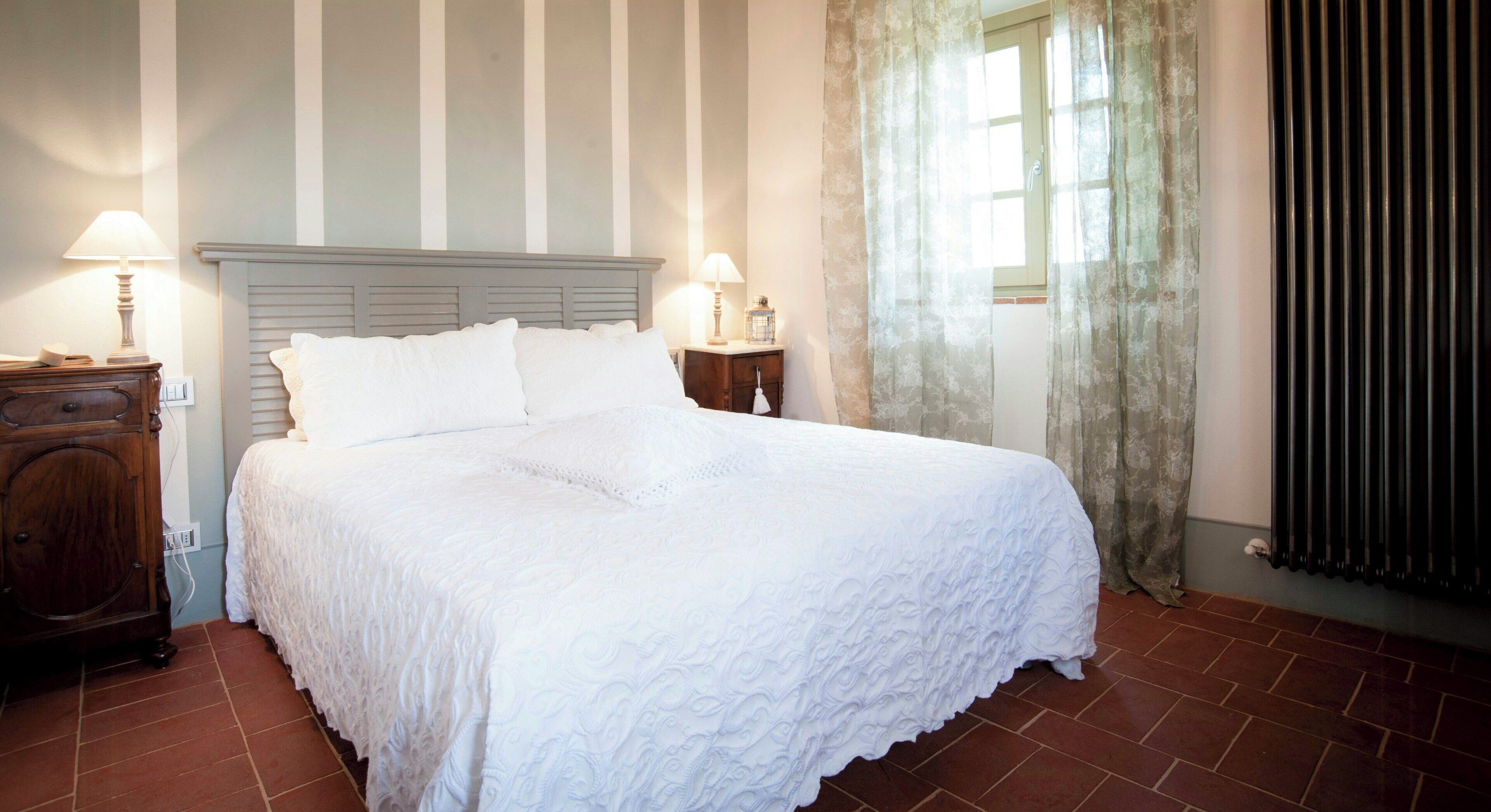Una cama bien hecha con una colcha blanca, dos lámparas de noche, una mesita de madera y una ventana con cortinas.