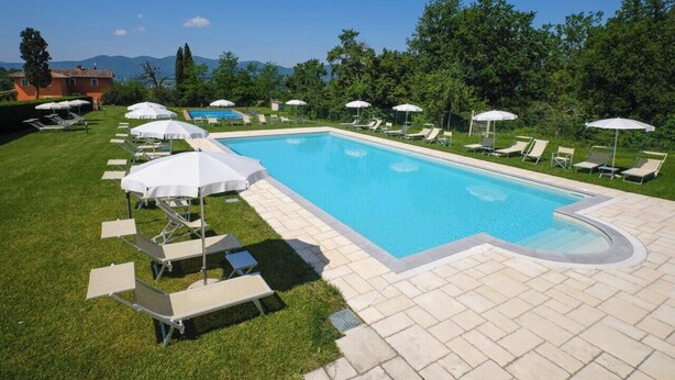 Piscina com espreguiçadeiras e guarda-sóis, cercada por gramado e árvores.