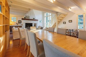 A spacious living area with a long wooden table, comfortable seating, a fireplace, and a staircase.