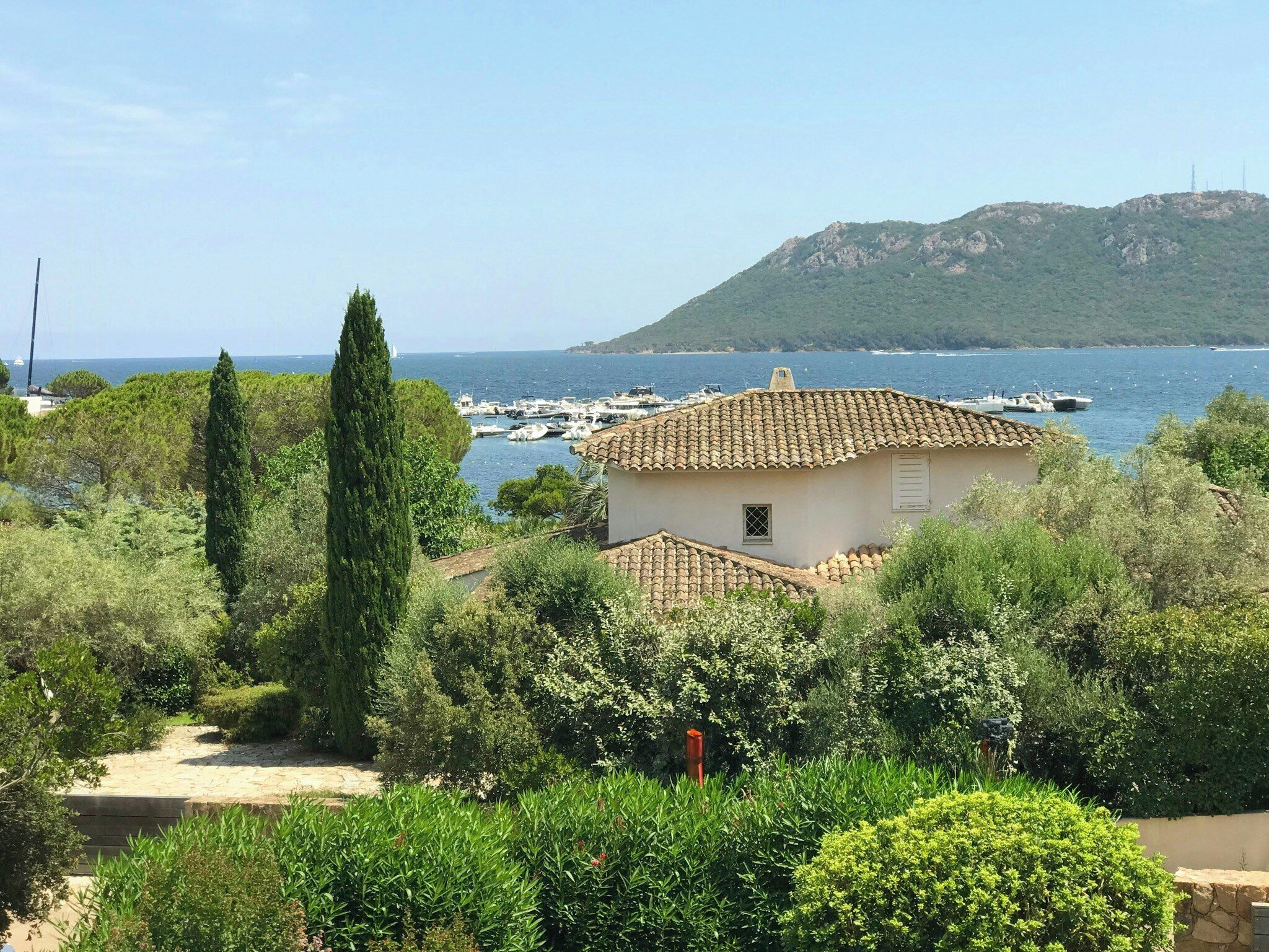 A coastal building with a tiled roof, surrounded by greenery and overlooking a marina with boats.