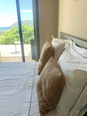 A bed with white bedding and two beige pillows, a window with a view of greenery and mountains, and a metal table in the foreground. - Villa with sea view and garden in Corsica (Porto-Vecchio)
