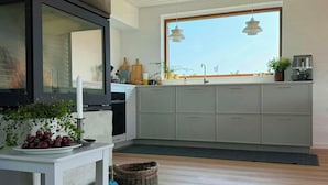 A modern kitchen with grey cabinets, a large window, and a dining area with a table and potted plants. - Cozy summer house in Agger near the North Sea (Vestervig)
