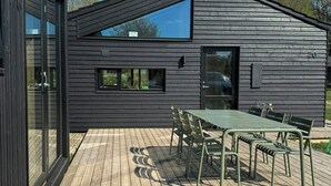 Een modern gebouw met een houten terras, een eethoek met stoelen en een tafel, en een groot raam.