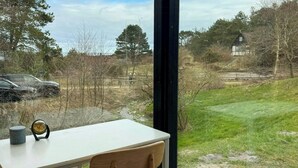 Een kamer met een bureau en stoel, grote ramen en uitzicht op een grasveld met bomen en een huis.