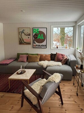 Una sala de estar con un sofá gris, una mesa de centro de madera y un sillón con un plaid suave. Hay dos carteles colgados en la pared y una ventana con vistas a árboles.