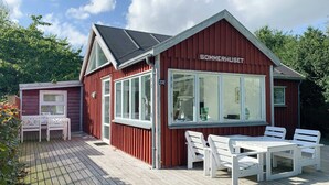 En röd träbyggnad med namnet ”SOMMERHUSET” på sidan, en terrass med vita möbler och en klar himmel.