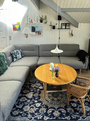 Ein modernes Wohnzimmer mit einem grauen Ecksofa, einem runden Holztisch, einem Korbstuhl und einem gemusterten Teppich.