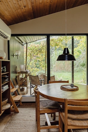 A wooden dining table with chairs, a hanging pendant light, and a view of the outdoors through large windows. - Stylish oasis in the middle of Kulhuse (Kulhuse)