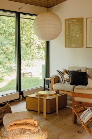 A modern living room with large windows, a sofa, a coffee table, and a hanging light fixture. - Stylish oasis in the middle of Kulhuse (Kulhuse)