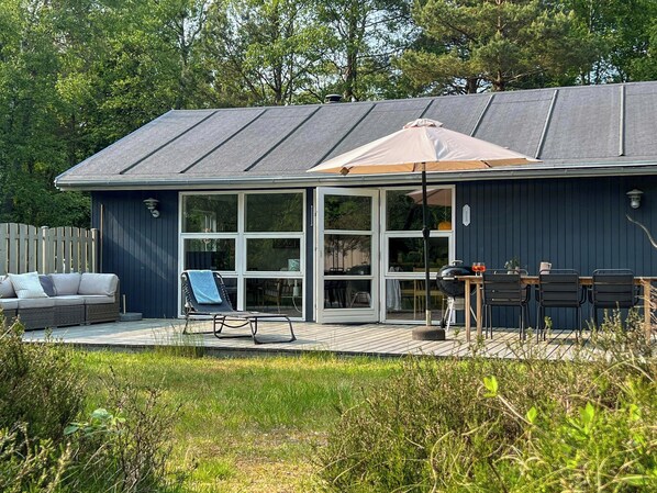 A house with a deck, a sofa, a lounge chair, and a table with chairs. - Renovated holiday home by forest lake in Dueodde (Nexø)