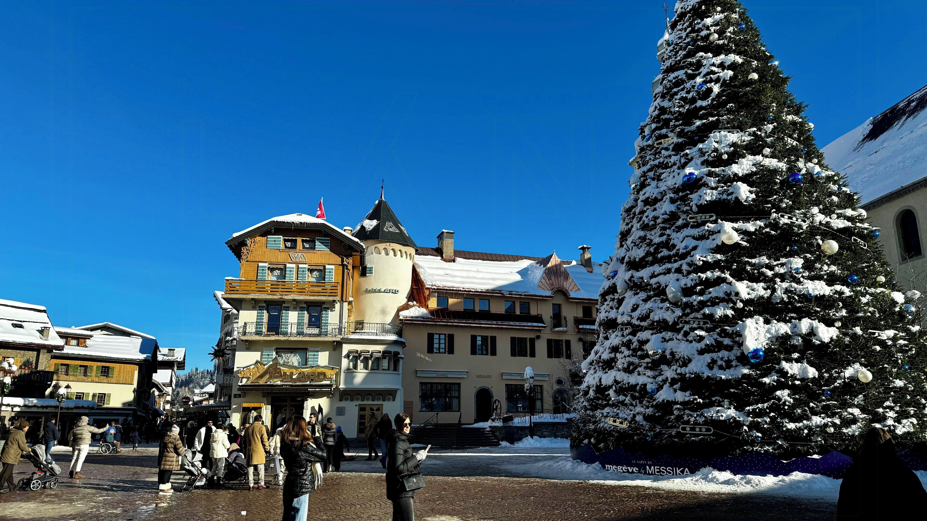 Un sapin de Noël recouvert de neige, installé sur une place de ville, avec des bâtiments historiques en arrière-plan.