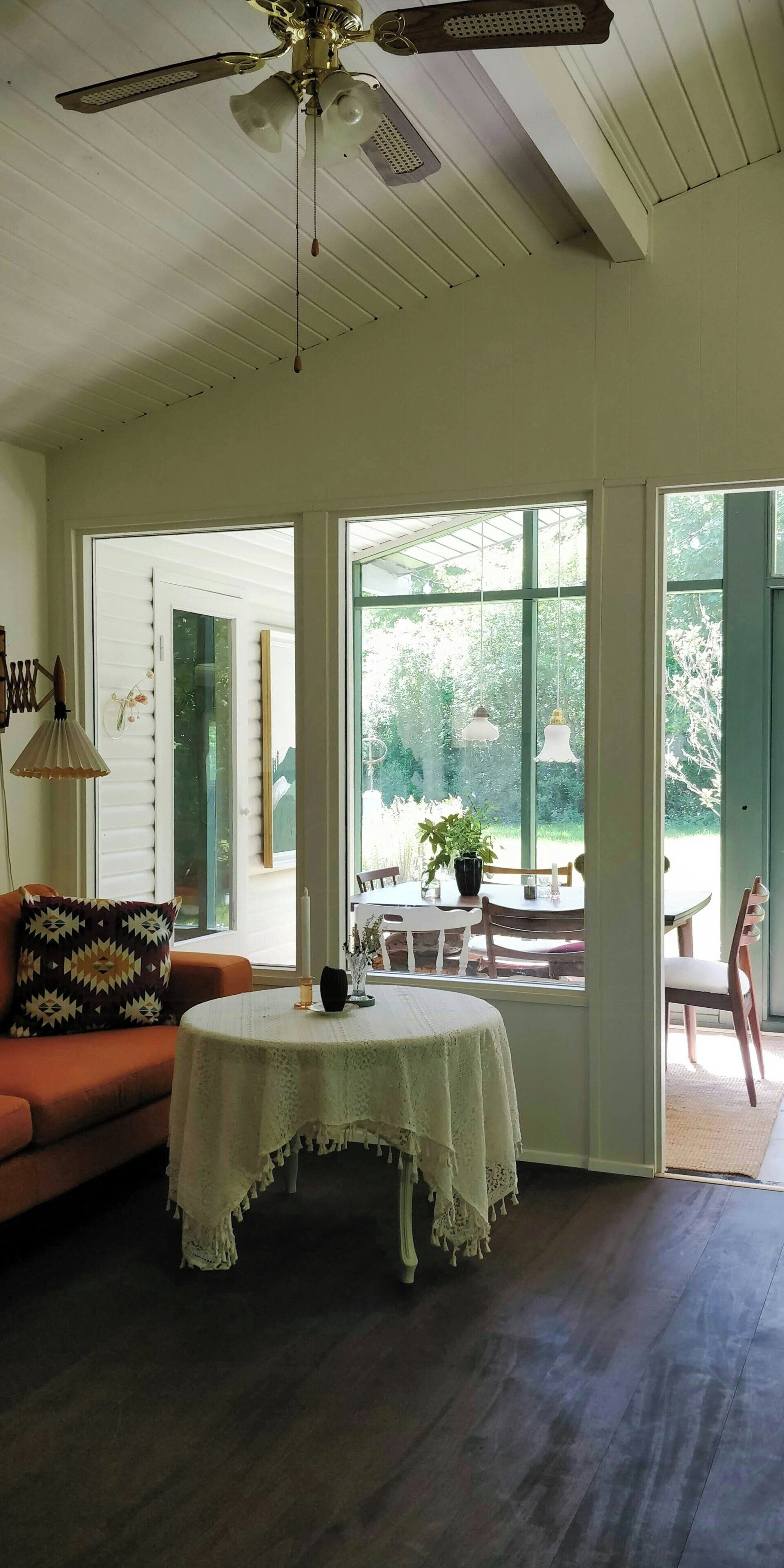 A room with a ceiling fan, a table with a lace tablecloth, a sofa with a patterned pillow, and large windows with a view of greenery.