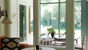 Quarto com ventoinha no teto, mesa com toalha de renda, sofá com almofada estampada e grandes janelas com vista para o verde.