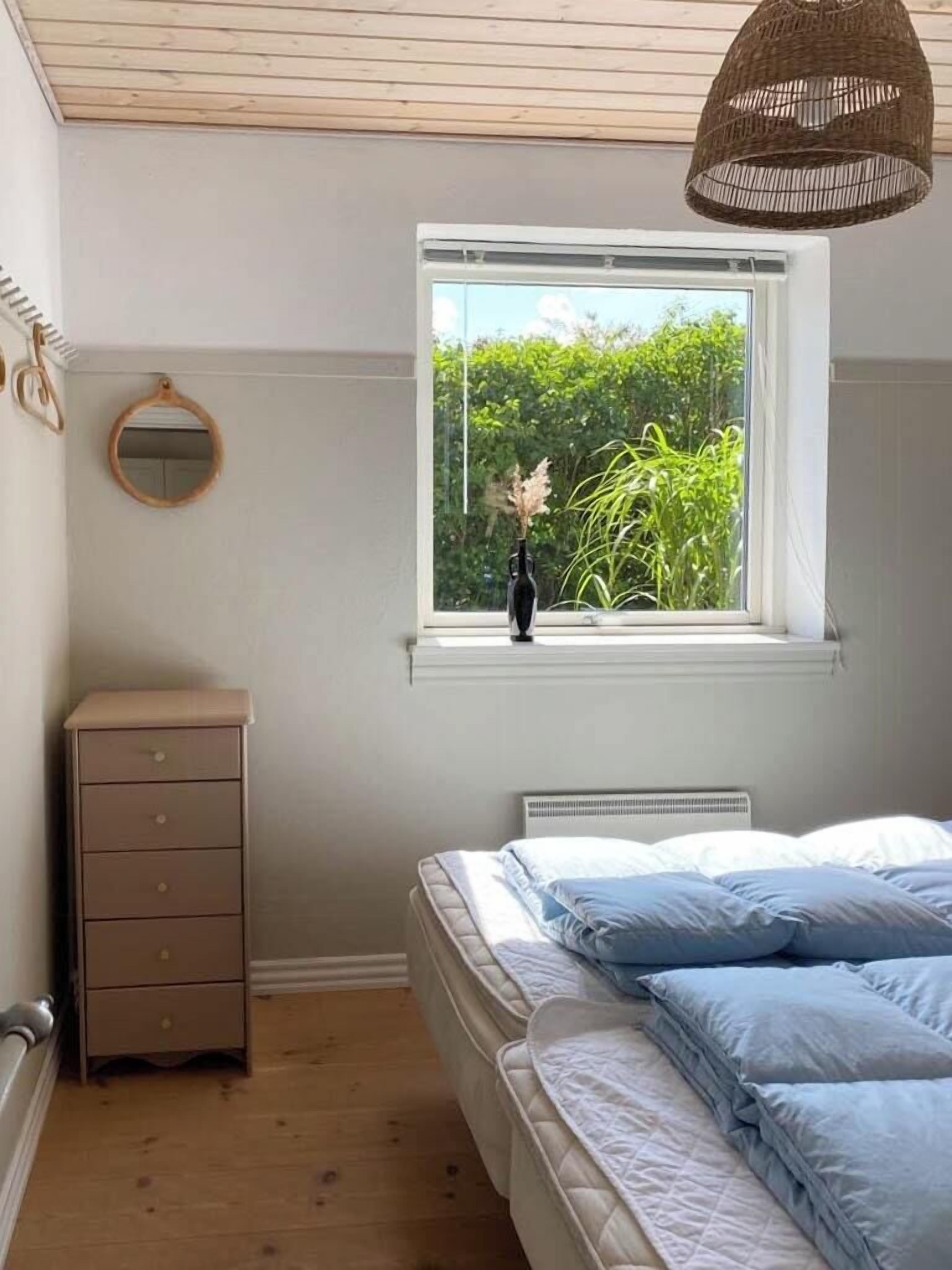 Un dormitorio con cama, cómoda, ventana con vista a la vegetación y lámpara colgante.