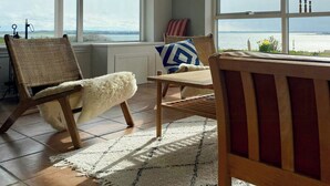 Une pièce dotée de grandes fenêtres, d’un fauteuil en osier, d’une table en bois et offrant une vue sur la mer.