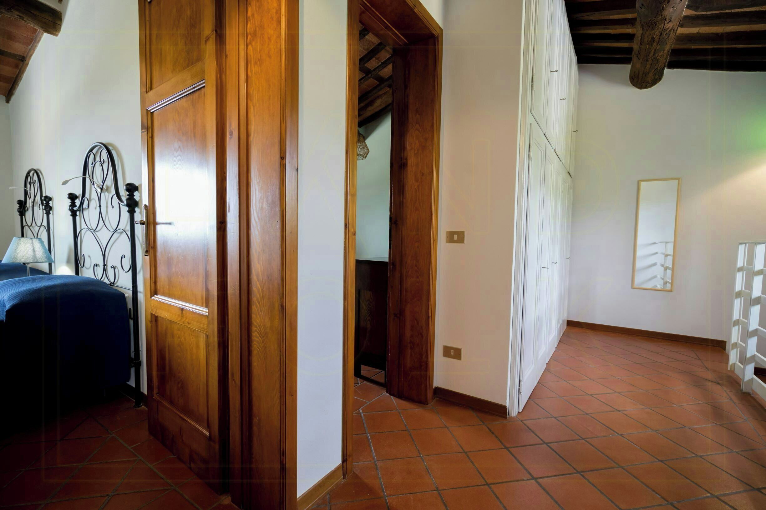 A room with a bed, wooden door, tiled floor, and a hallway leading to another room.